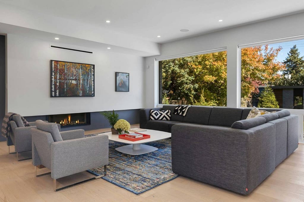 Contemporary living room with gray sectional, patterned chairs, fireplace, blue rug, coffee table, and large windows overlooking colorful autumn trees.