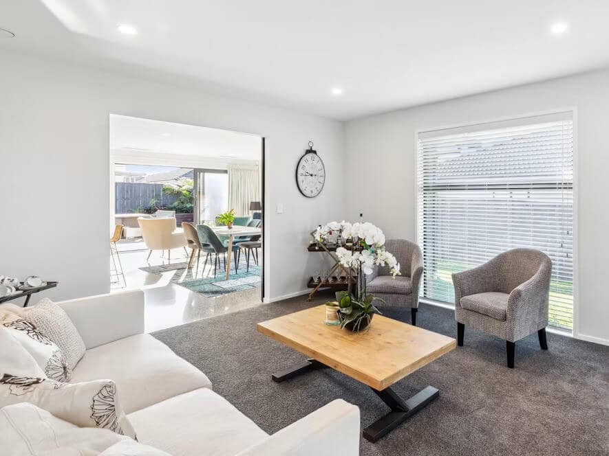 Bright, modern living room with white sofa, wood coffee table, flowers, gray armchair, large windows, and adjoining dining area; calm, inviting mood.