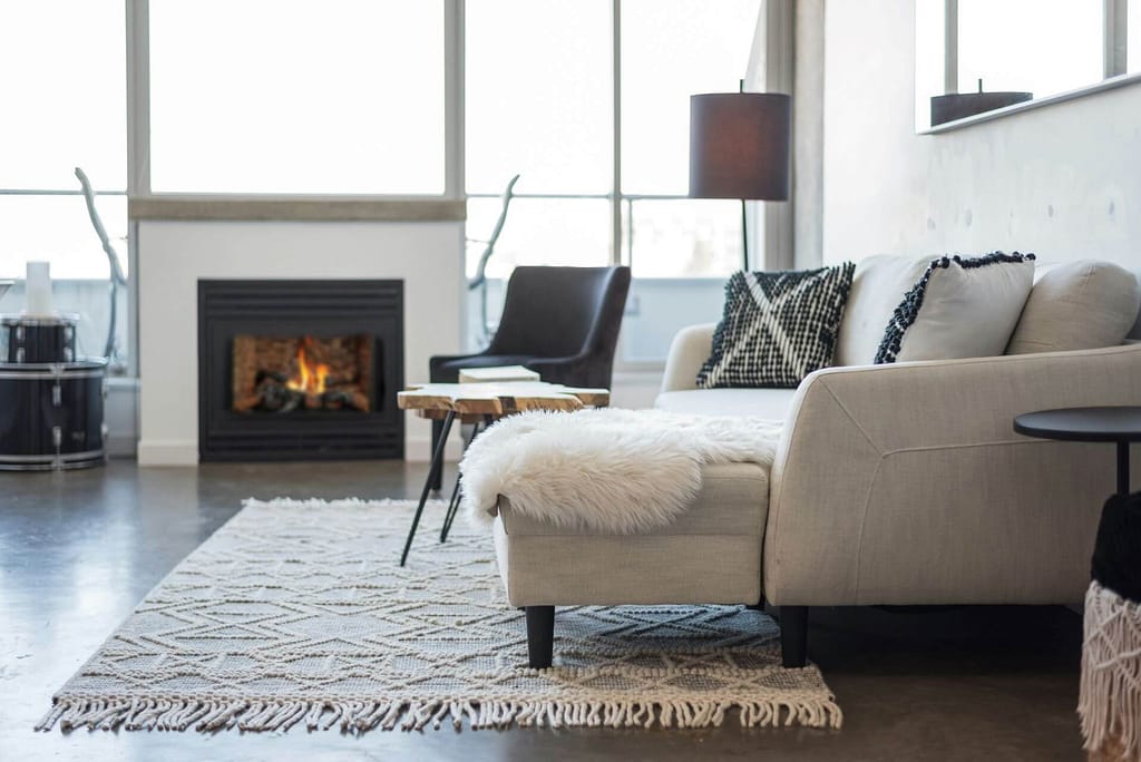 Cozy modern living room with a lit fireplace, beige sectional sofa, textured rug, plants, and large windows creating a warm, inviting mood.