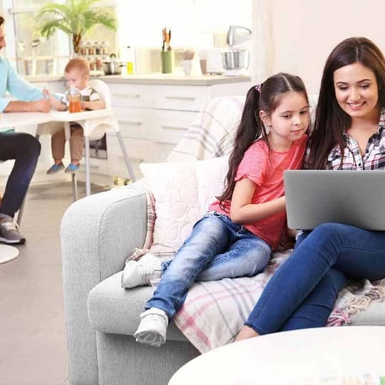 EVOAQ Home Ventilation image mother with daughter relaxing on couch on right looking at laptop father feeding baby son in background